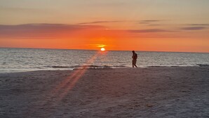 Escena de playa con atardecer, una persona de pie sobre la arena y el océano al fondo.