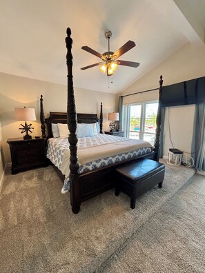 A four-poster bed with a ceiling fan, a bedside table with a lamp, and a view of the outdoors through a window.