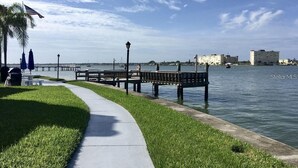 Un paseo marítimo con un muelle de madera que se adentra en el agua, una zona verde y un cielo despejado.
