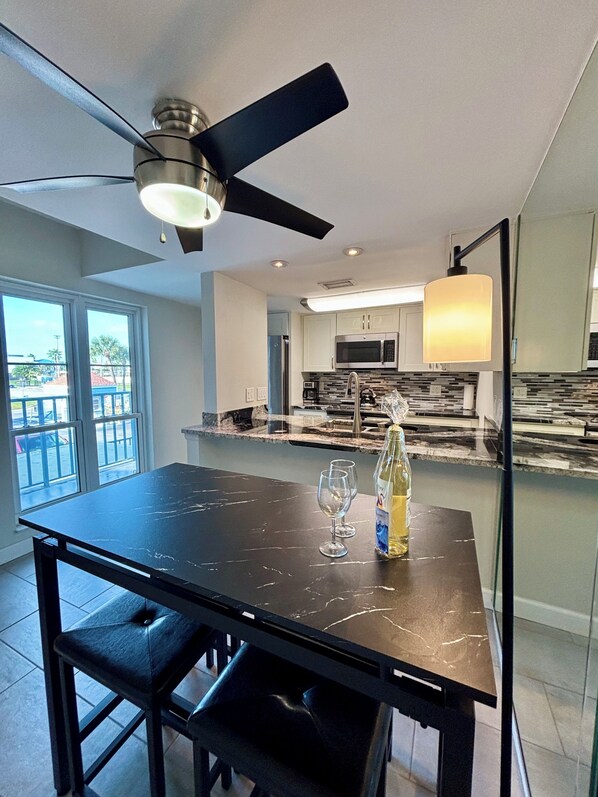 A modern kitchen with a dining area, featuring a black marble table, two wine glasses, a bottle of wine, and a ceiling fan.