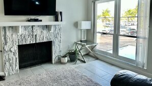 Una sala de estar moderna con televisión de pantalla plana, chimenea, un ventanal con vista a un estacionamiento y una pequeña mesita de noche con lámpara.