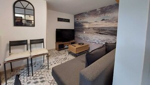 Una sala de estar con un televisor grande, dos sillones, un sofá y un mural de playa, además de un letrero que dice "SEA FOOD MARKET".