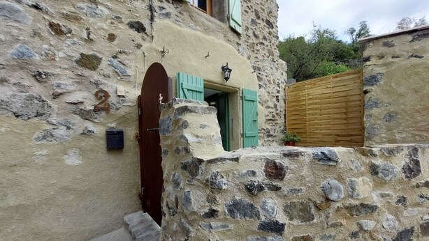 Un edificio rústico de piedra con puerta de madera y contraventanas verdes, una pequeña ventana con farol y una valla de madera.
