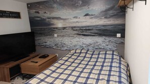 Un dormitorio con una cama, televisión, escritorio y un mural en la pared que representa una playa.