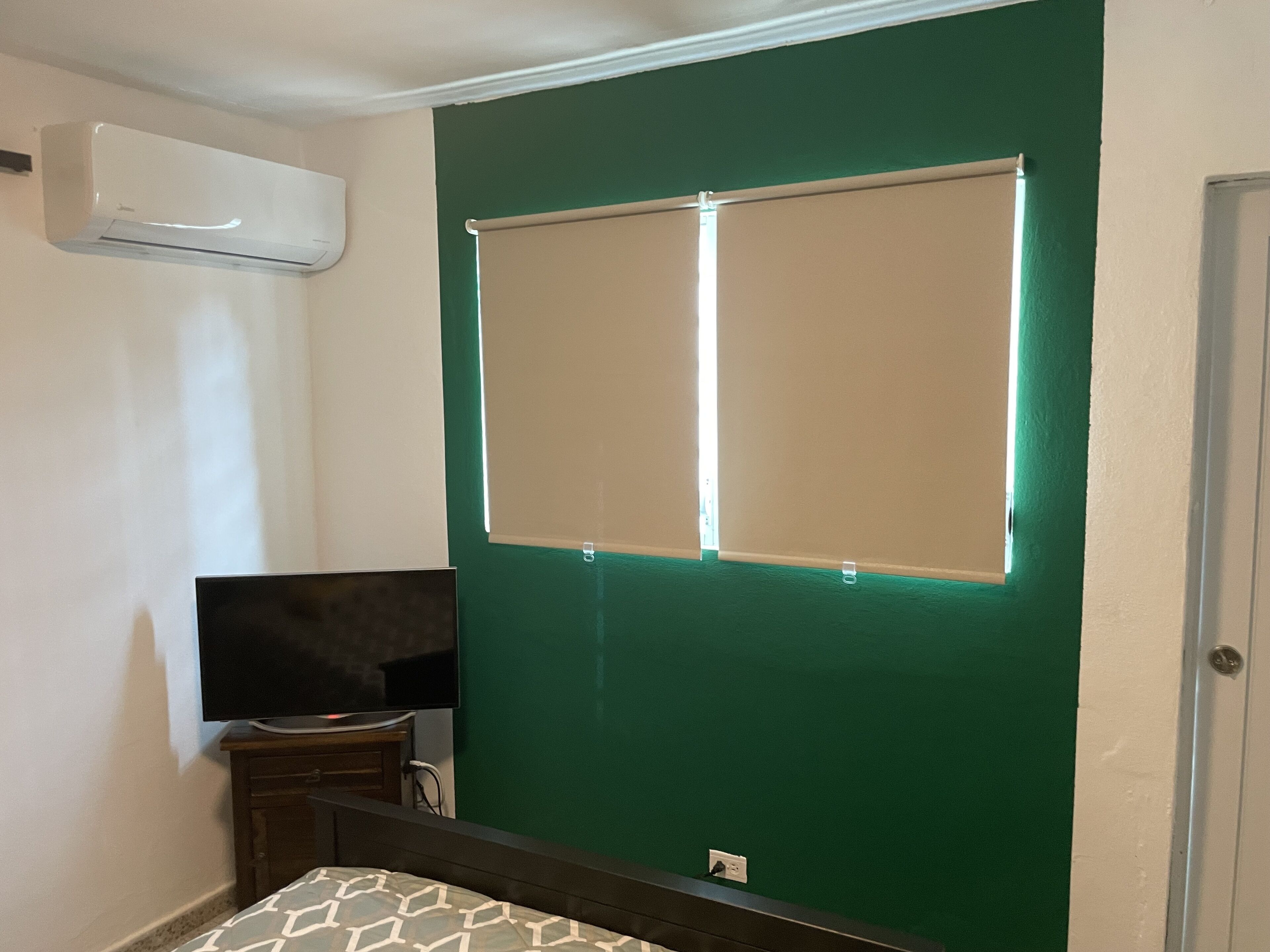 A room with a green wall, a flat-screen TV on a wooden stand, a bed with a patterned bedspread, and an air conditioning unit mounted on the wall.
