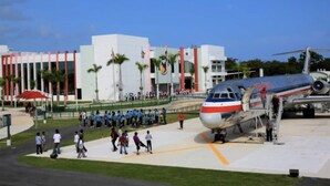 An airport terminal with a large airplane parked in the foreground, a group of people gathered near the terminal entrance, and a building with a modern design in the background. - studio at carolina (Carolina)