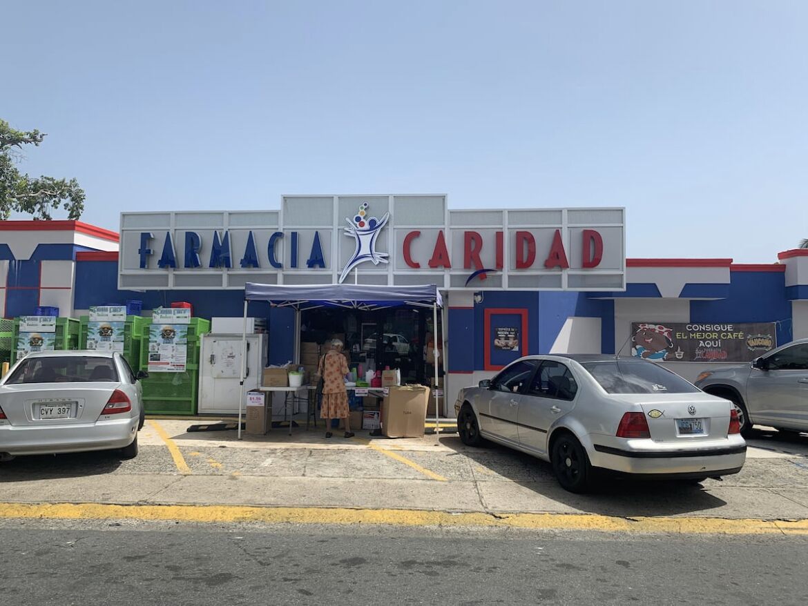 A pharmacy named 'Farmacia Caridad' with a blue and white facade, a storefront, and a parking lot with cars.