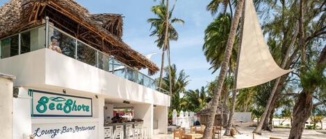 A beachside lounge-bar restaurant with a thatched roof, outdoor seating, and palm trees.