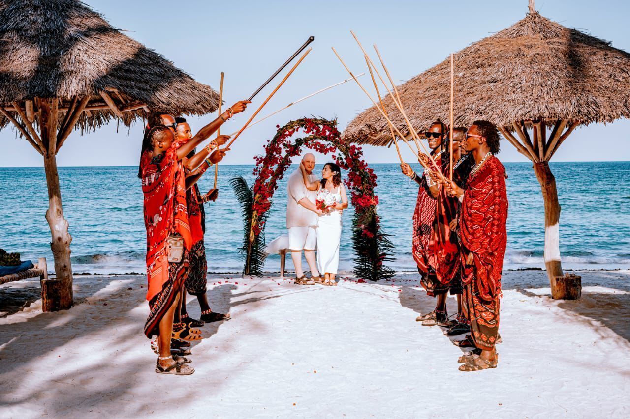 Une cérémonie de mariage sur la plage, avec une arche nuptiale blanche et des invités tenant des bâtons.