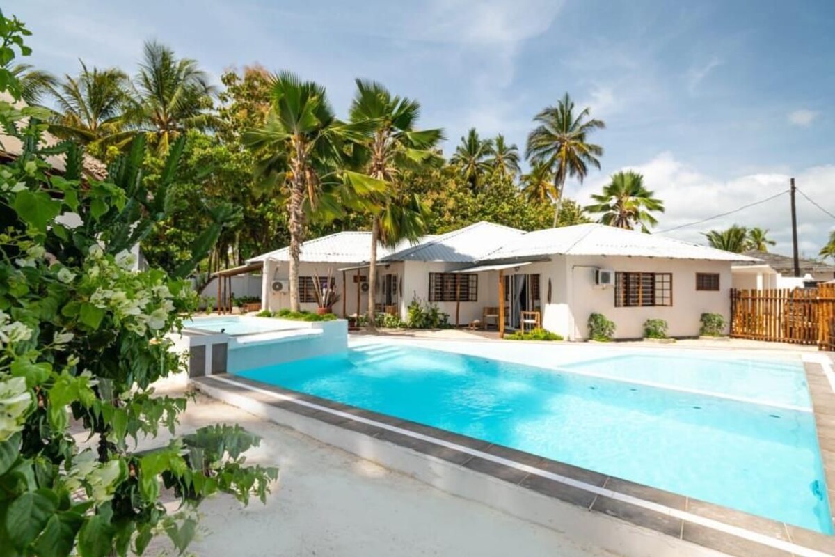 Una piscina con plataforma blanca, rodeada de palmeras y un edificio con techo blanco.