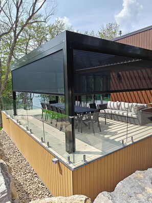 A modern outdoor dining area with glass railings, a wooden deck, and a view of the water.