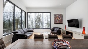 A modern living room with a large window, a black leather sofa, a dining table, and a flat-screen TV mounted on the wall.