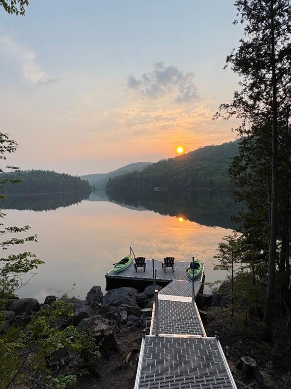 A dock with chairs and a kayak, overlooking a calm lake with a sunset in the distance.