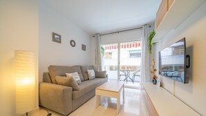 A modern living room with a sofa, a coffee table, a TV mounted on the wall, and a balcony with a view of the street.
