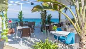 An outdoor seating area with tables and chairs, surrounded by potted plants and a view of the sea.