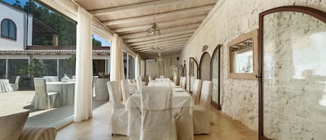 A long dining room with white tablecloths and chairs, a wooden ceiling, and large windows.