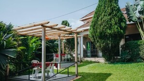 A covered outdoor seating area with a table and chairs, surrounded by greenery and a tall tree.