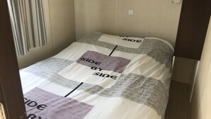 Ett litet, smalt rum med en säng som har ett mönstrat överkast med orden ”SIDE BY SIDE” upprepade. Det finns två väggmonterade ljusströmbrytare och ett fönster med vertikala persienner.