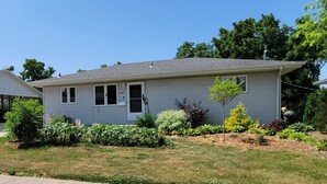 A single-story house with a grey exterior, a small front yard with various plants, and a clear blue sky. - Private entrance/space. Peaceful getaway. Close to events. King bed. 
Amenities! (Dubuque)