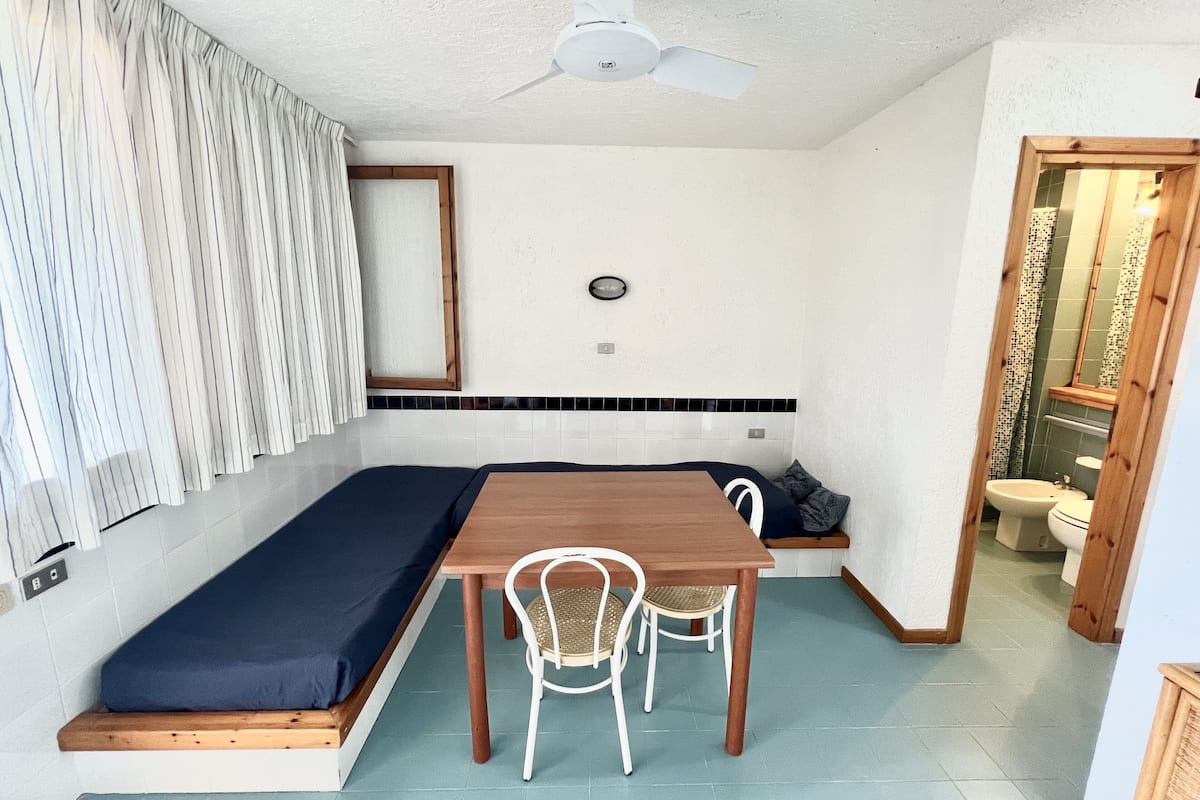 A compact room with a bed, a small table, and chairs. There is a ceiling fan, a window with curtains, and a bathroom visible through an open door.
