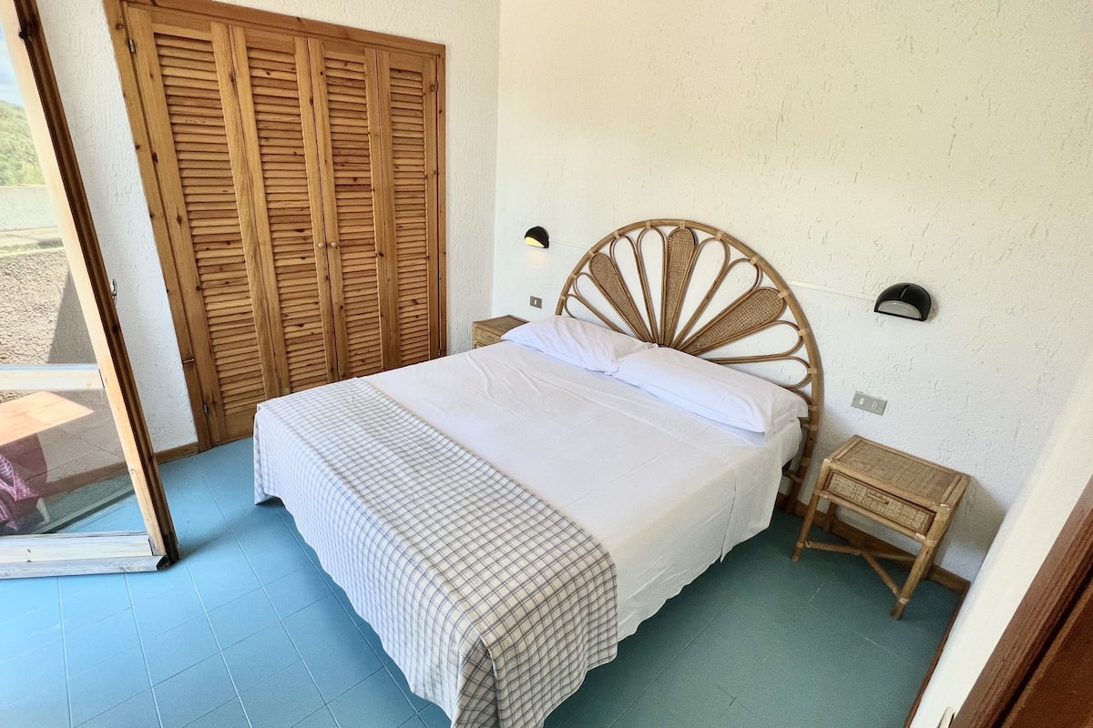 A bedroom with a bed, a wooden wardrobe, a small bedside table, and a wall-mounted light fixture.