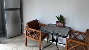 Un espacio habitable compacto con kitchenette, que incluye un refrigerador, una pequeña mesa con sillas y una planta en maceta.