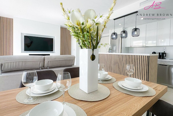 Et moderne spiseområde med et træbord dækket til fire personer, en hvid vase med blomster og et fjernsyn i baggrunden.