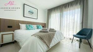 Ein modernes Hotelzimmer mit einem großen Bett, einem Nachttisch, einem Sessel und einem Fenster mit durchsichtigen Vorhängen.