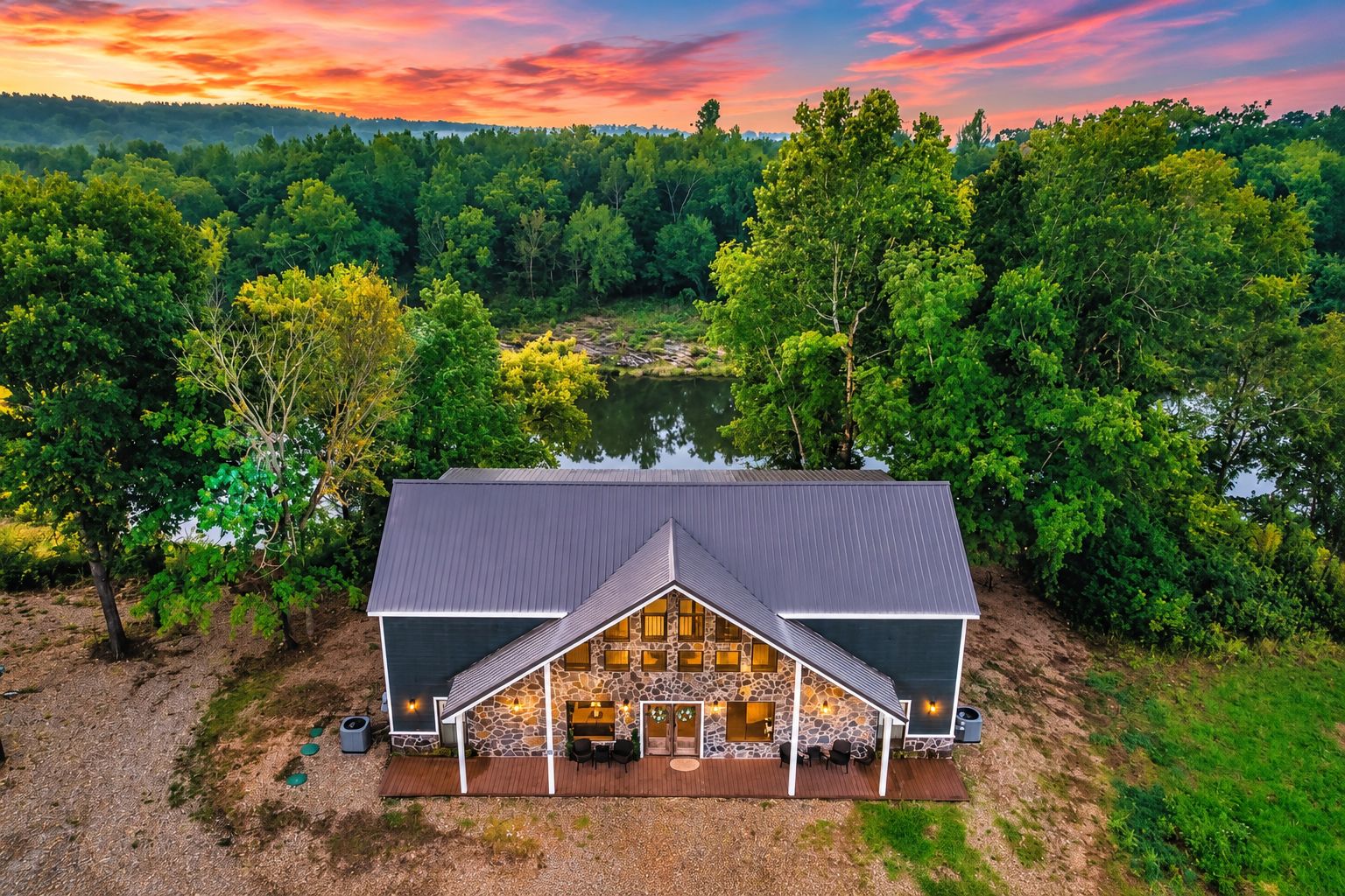 A two-story stone building with a sloped roof, a patio with outdoor seating, and a view of a lake surrounded by trees.