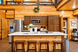 A wooden cabin interior with a large kitchen island, bar stools, a refrigerator, a microwave, and a sink.