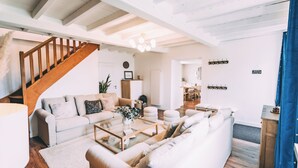 Een ruime woonkamer met een hoge plafond, een houten trap, een bank, een salontafel en een eethoek.