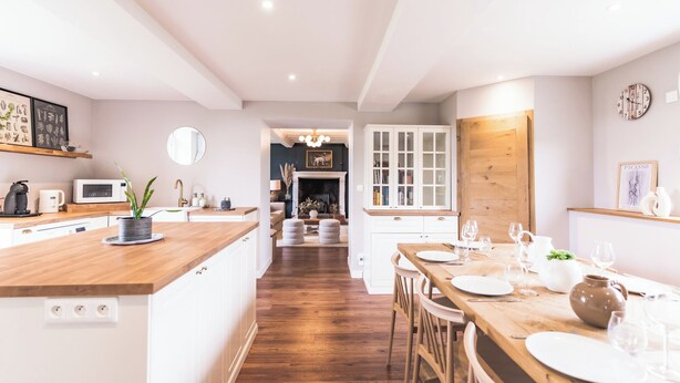 Een moderne keuken met houten aanrechtbladen, witte kasten en een centraal eiland. Er is een eethoek met een houten tafel gedekt voor zes personen, en een open haard op de achtergrond.