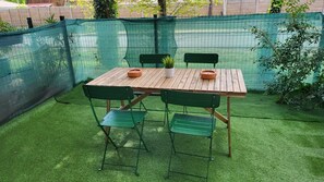 A wooden table with four green chairs, surrounded by artificial grass, with a green mesh fence in the background.