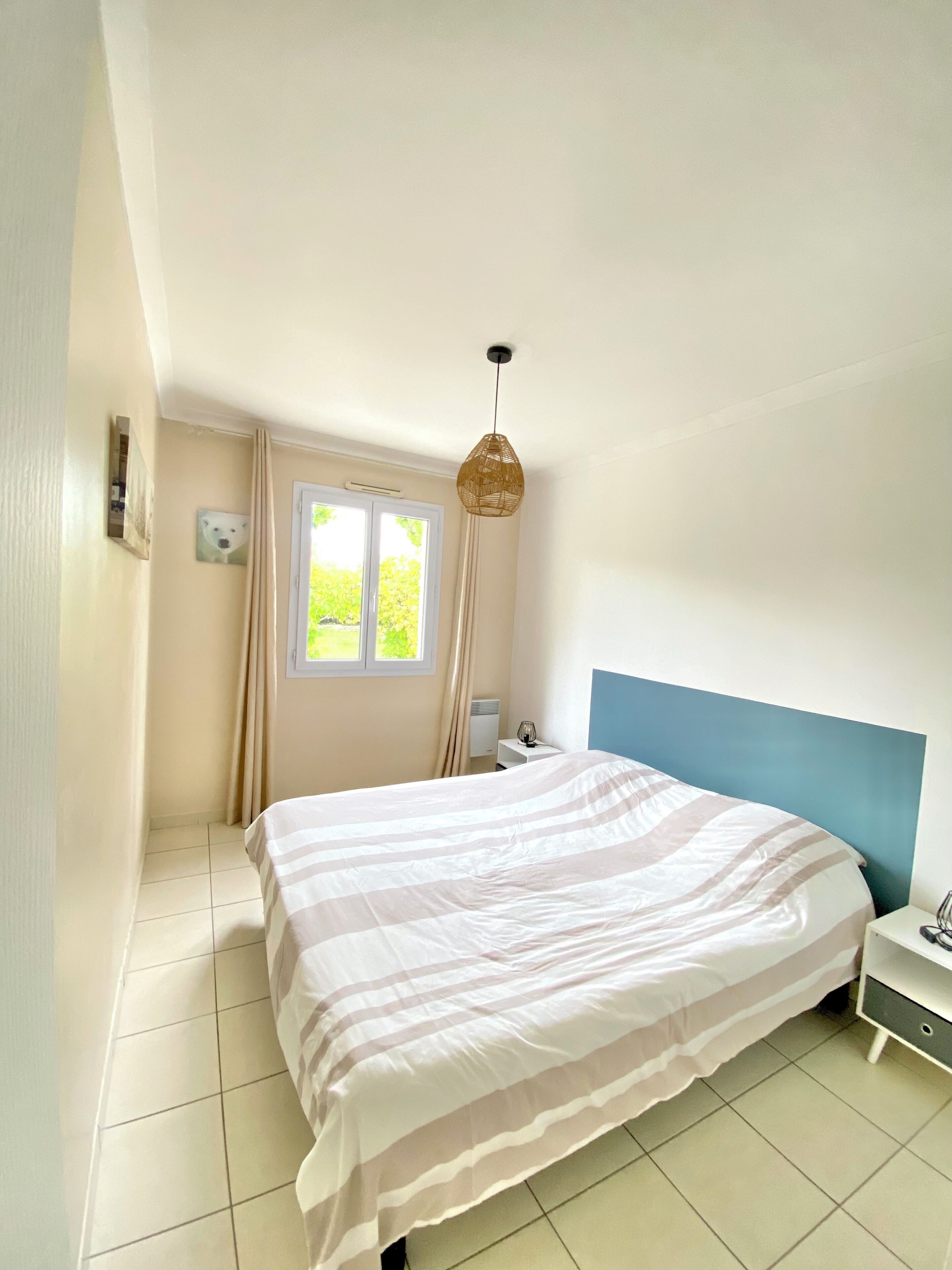 Une chambre à coucher comprenant un lit, une table de chevet, une commode, une fenêtre et un luminaire suspendu.