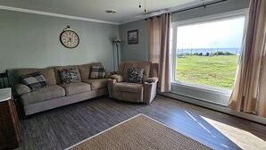 A living room with a sofa, armchair, and a large window offering a view of a grassy area. - New Waterfront! Drop Anchor on Brannocks Bay. An Amazing waterfront retreat! (Cambridge)