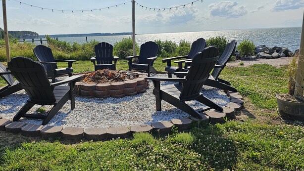 A circular seating arrangement with four Adirondack chairs around a fire pit, set on a gravel area with a view of a lake and distant shoreline. - New Waterfront! Drop Anchor on Brannocks Bay. An Amazing waterfront retreat! (Cambridge)