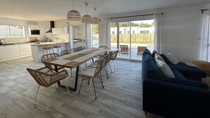 Moderni keittiö, jossa on valkoiset kaapit, ruokailutila pitkällä puupöydällä ja rottinkituoleilla sekä sininen sohva olohuoneessa, josta on liukuovet takapihalle.