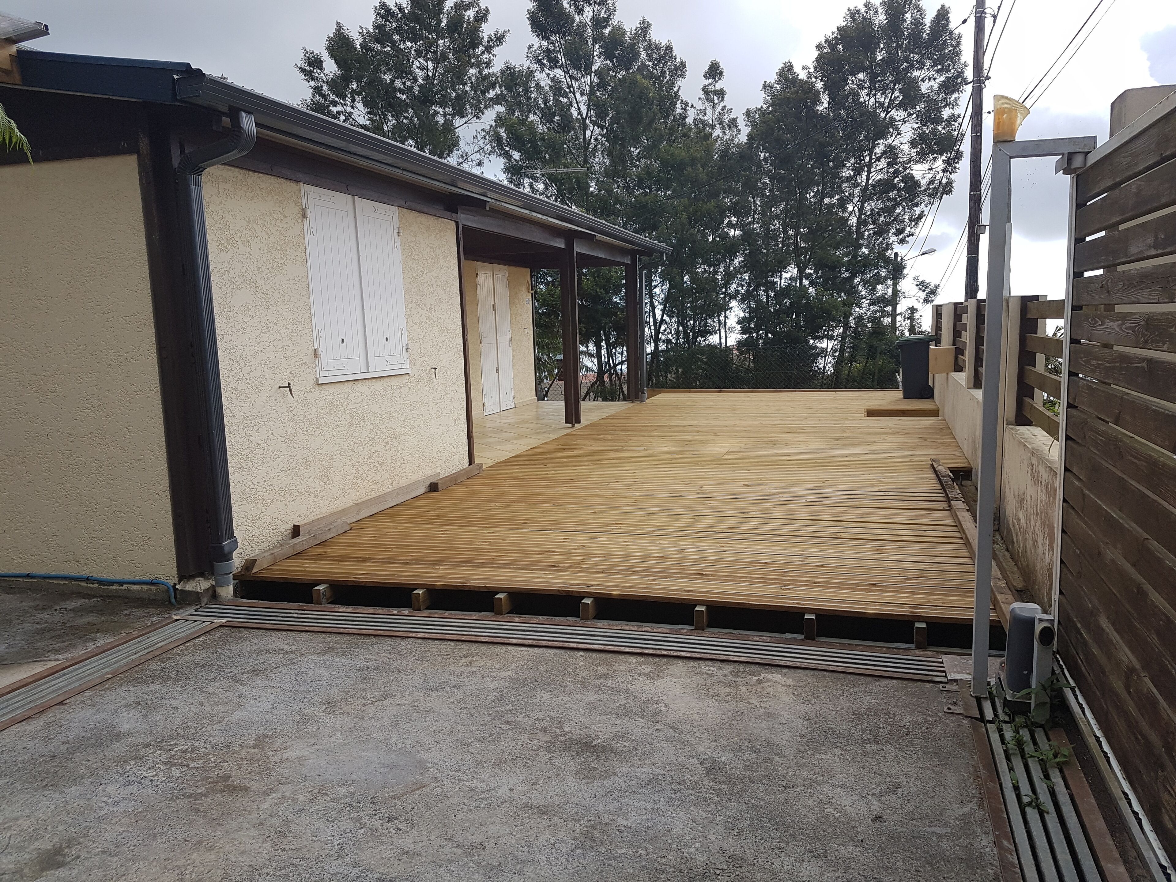 A covered wooden deck with a metal railing, a concrete floor, and a beige wall with a window.