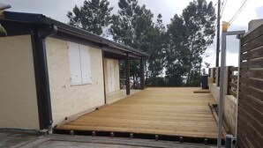 Een overdekt houten terras met een metalen hekwerk, een betonnen vloer en een beige muur met een raam.