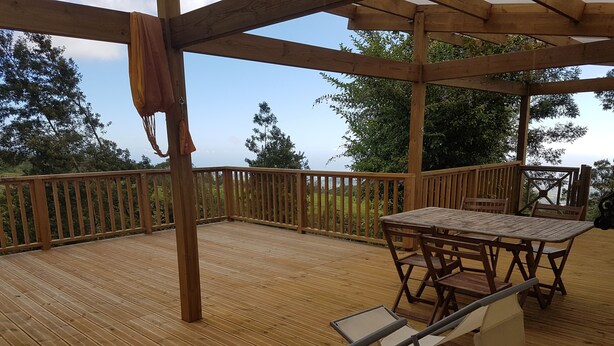 Een houten terras met een tafel en stoelen, omgeven door een houten balustrade en bomen.