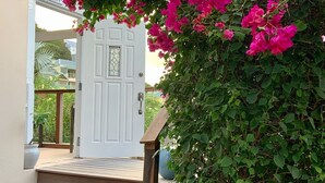 Uma porta branca com um painel de vidro, rodeada por folhagem verde exuberante e flores rosa vibrantes.