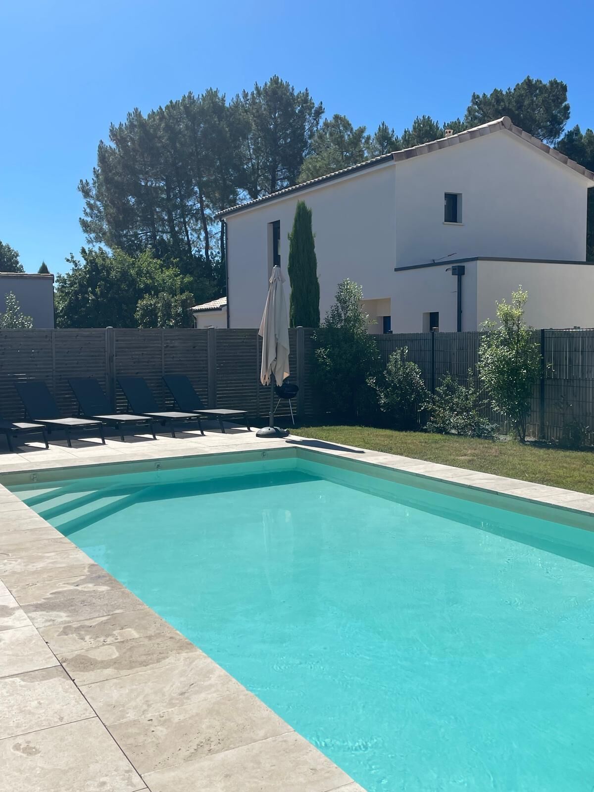 Une piscine rectangulaire, bordée d’une terrasse carrelée, entourée d’une clôture en bois et d’un bâtiment blanc doté d’un toit plat.