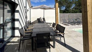 Een terras buiten met een houten pergola, een lange tafel met stoelen en een zwembad op de achtergrond.