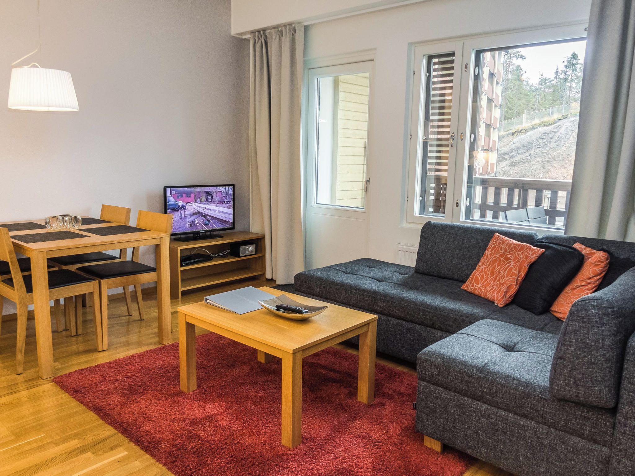 Kalustettu olohuone, jossa on sohva, ruokapöytä, tuoleja, televisio ja ikkuna, josta avautuu näkymä rakennukseen ja puihin.