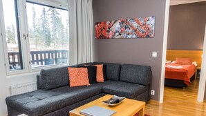 Un salon moderne avec un canapé d’angle gris, une table basse en bois et un tapis rouge. Un tableau orne le mur, et une fenêtre offre une vue sur les arbres.