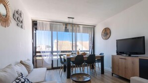 Un salon moderne comprenant un canapé, une table à manger avec des chaises, un téléviseur posé sur un meuble en bois et une grande fenêtre donnant sur des immeubles.