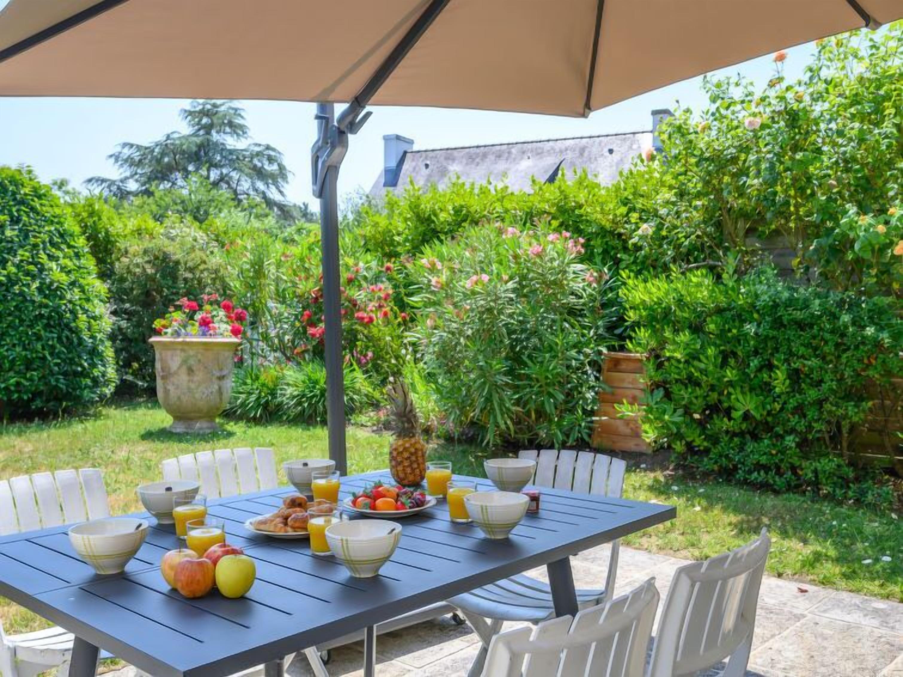 An outdoor dining area with a table set for breakfast, surrounded by lush greenery and a large planter with flowers.