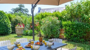 Un espacio al aire libre con zona de comedor, una mesa lista para el desayuno, rodeado de exuberante vegetación y una maceta grande con flores.