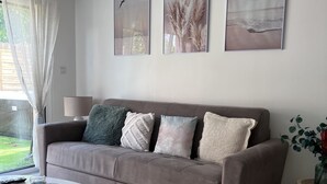 Ein Wohnzimmer mit grauem Sofa, drei Kissen, einem weißen Couchtisch und einem Teppich mit Muster. An der Wand hängen drei gerahmte Bilder und ein Fenster mit Vorhängen.