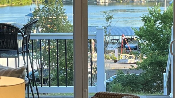 A balcony with a wicker chair and a table, overlooking a lake with a bridge and buildings in the background.